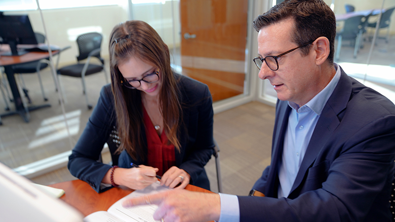 Businesswoman looks over papers while talking to a businessman points to a monitor