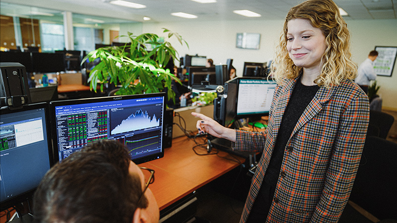 Business woman in sport coat points to a monitor of graphs while talking to business man 