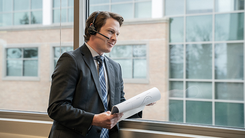Businessman talks to someone on a headset standing in front of windows looking out over another building