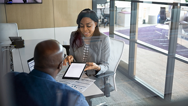Business woman points to a document on a tablet to a man sitting at a table with her in an office