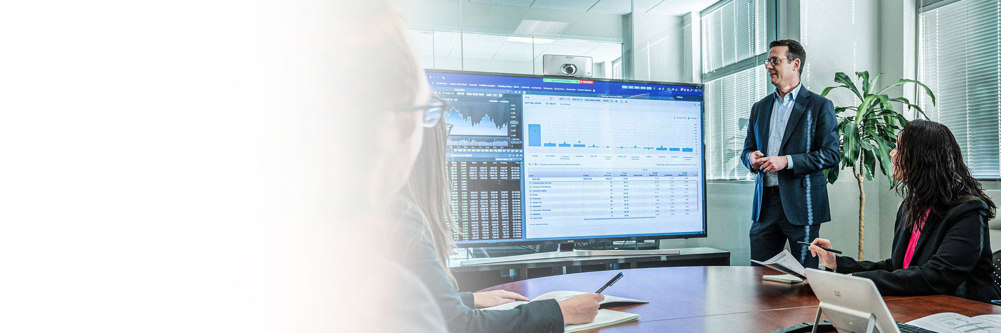 Business man conducts a meeting in front of a large monitor showing graphs