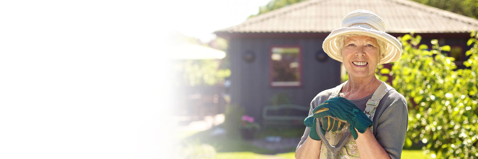 Woman in overalls and gardening hat leans against shovel smiling at camera with her purple house and green plants behind her