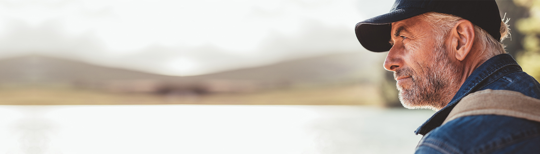 Man sitting at a lake wearing a baseball cap and hillside across the lake