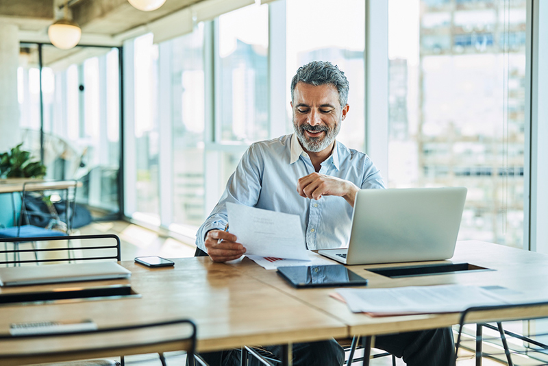 Man looks over a financial sheet while sitting in an office with his laptop open