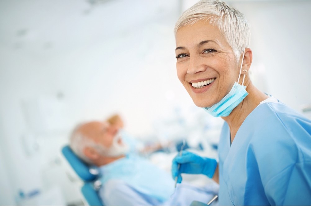 Woman dentist smiles at the camera wearing a blue smock while a man lays behind her in a dentist chair