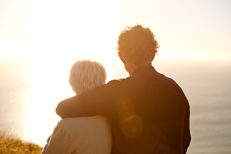 Man with arm around woman looking out over the ocean and the sunsetting in front of them