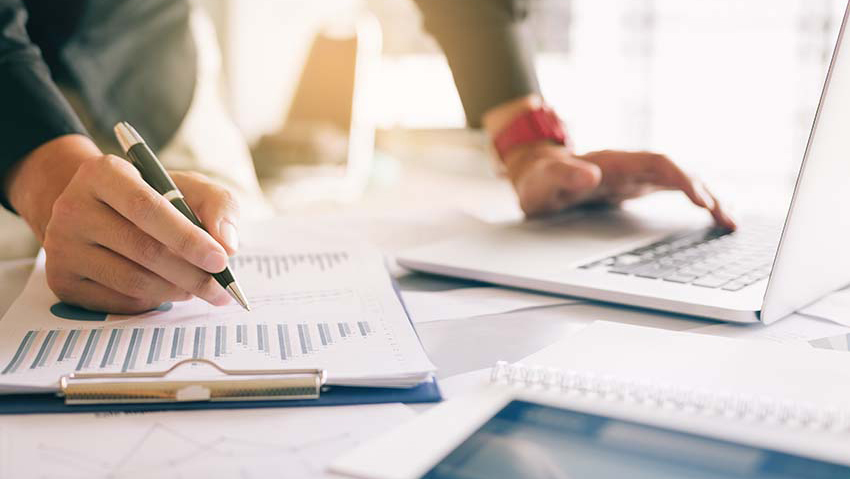 business professional working on a laptop with one hand and reviewing a graphs on a clipboard