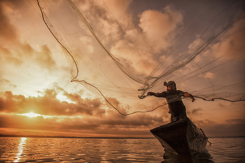 Man on a boat at sunset throws out a large fish net