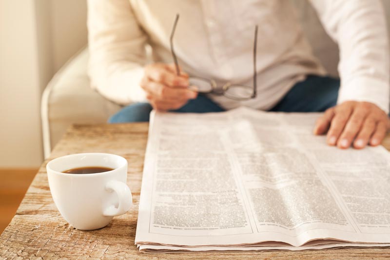 Man in white button up shirt holding glasses and looking at a newspaper on a coffee table with a cup of coffee on the table