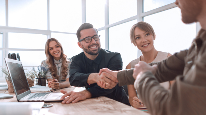 business professionals at a table around a laptop with two people shaking hands