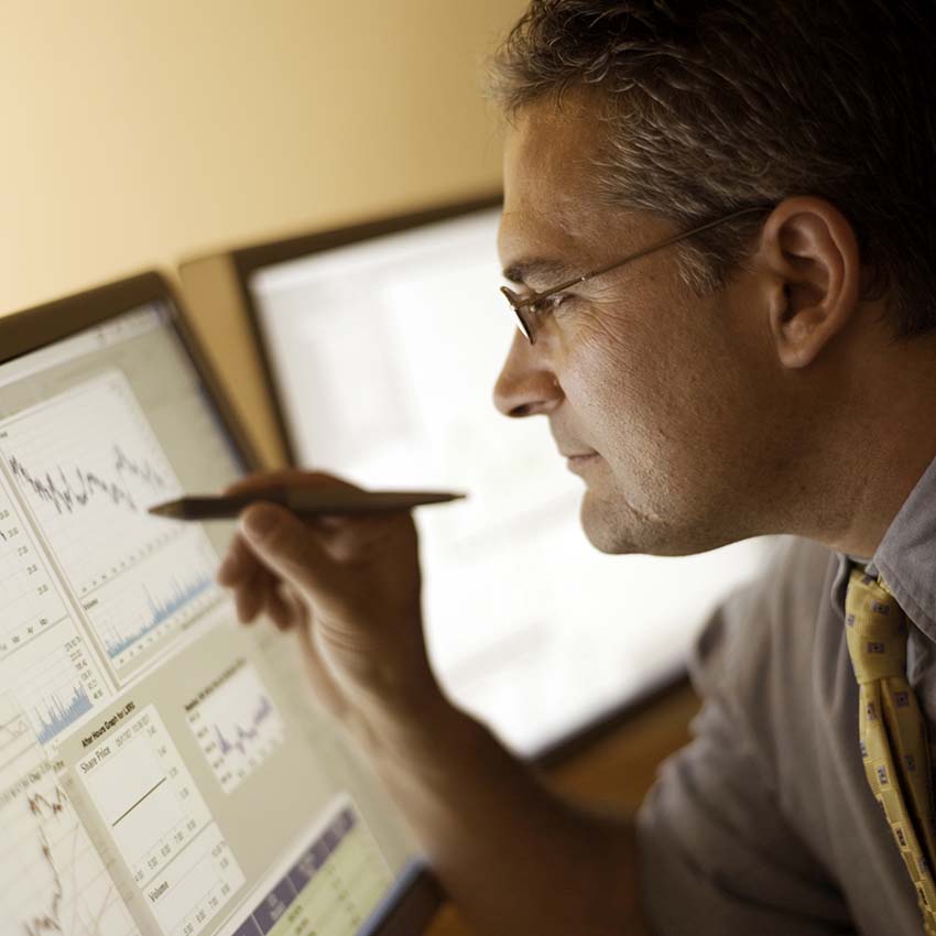 A business man holds his pen above graphs being displayed on monitors in front of him