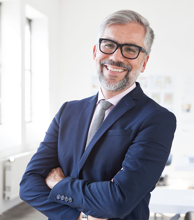 Man in blue suit smiles at camera with arms crossed standing in white office with windows