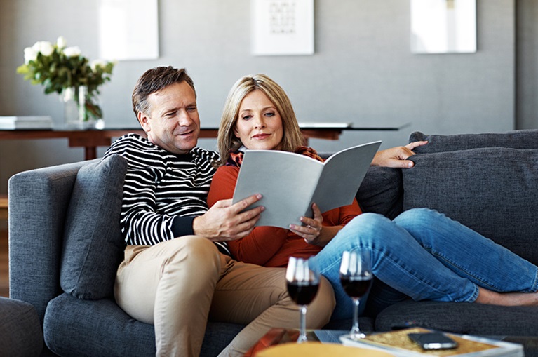 Man and woman sit on blue couch looking over a document with two glasses of wine on the coffee table