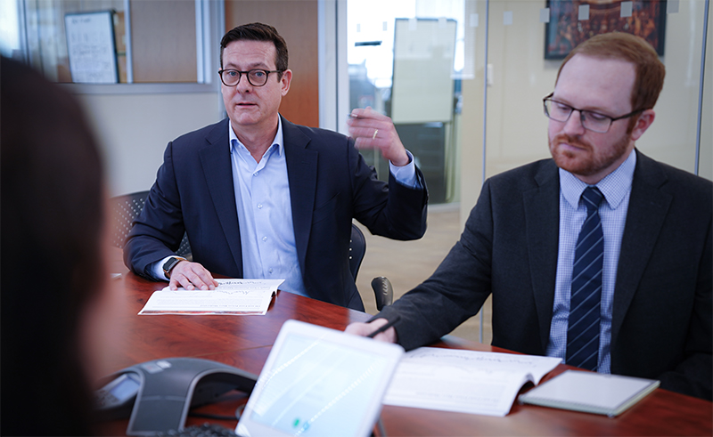 Two Men with glasses in suits talk with people at a table with papers on it
