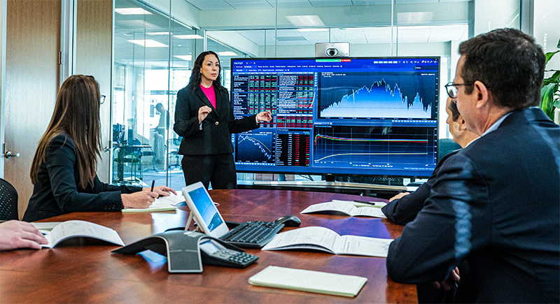Business woman conducts meeting in front of a large monitor with graphs and stocks