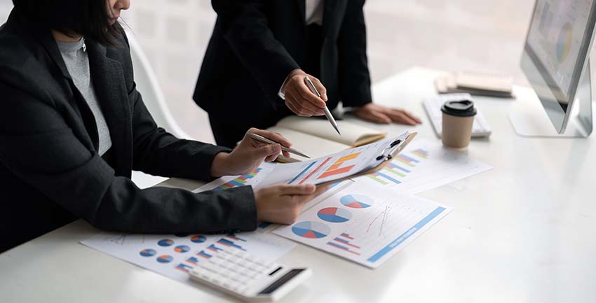 Woman looking at print outs of graphs spread out on desk while another woman points at a graph on a clipboard with a pen