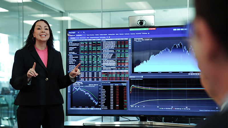 A business woman talking with her hands to a group, stands in front of a monitor displaying stock tickers and graphs 